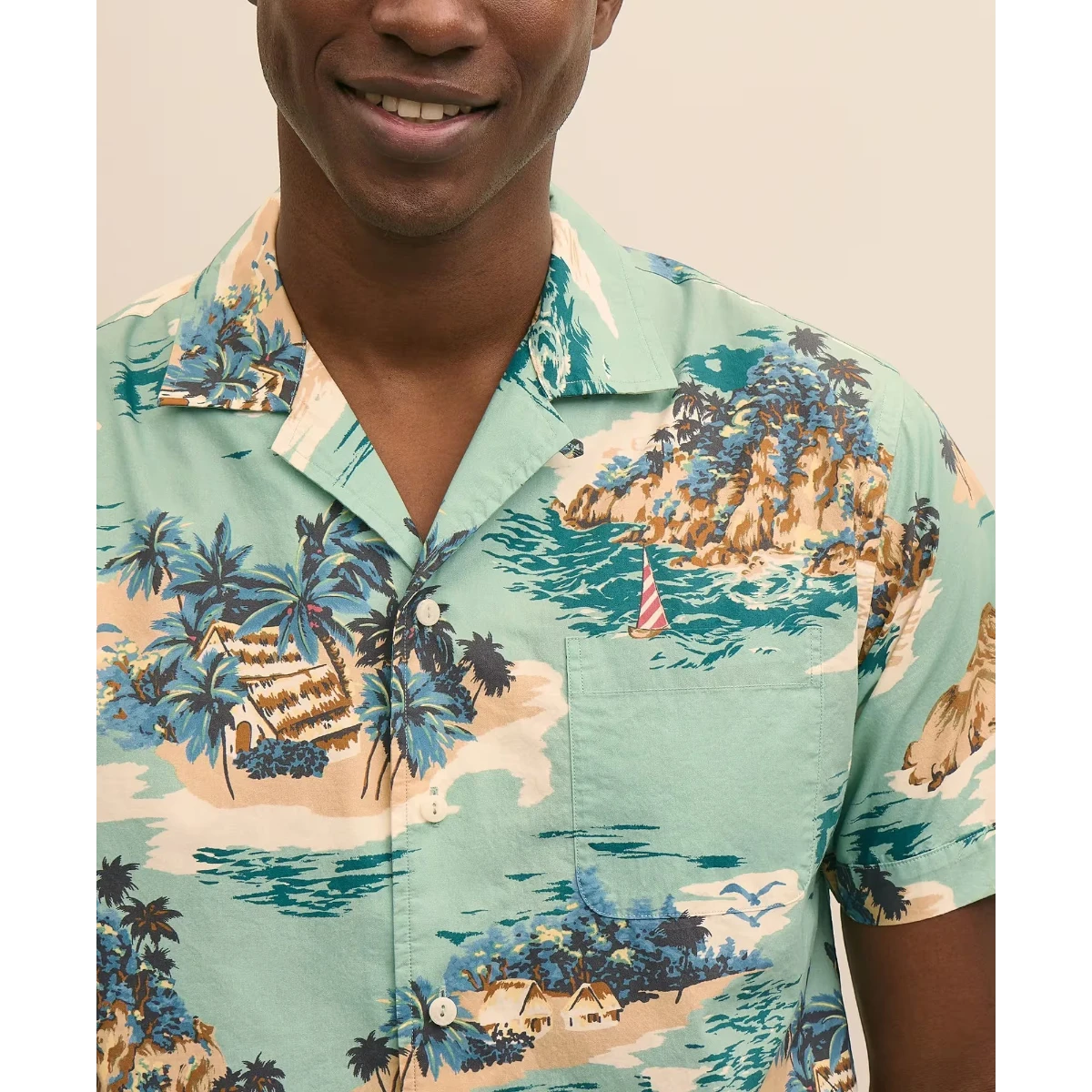 MILWORK blue 100% cotton camp collar shirt with an island motif pattern, featuring palm trees, huts, and a sailboat. Model is 6'0" and wears a size M.