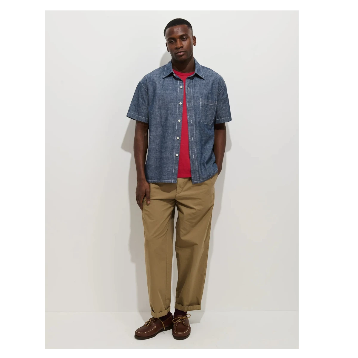 MILWORK Franklin short sleeve chambray shirt in indigo blue, worn over a red t-shirt, with tan trousers and brown boat shoes.