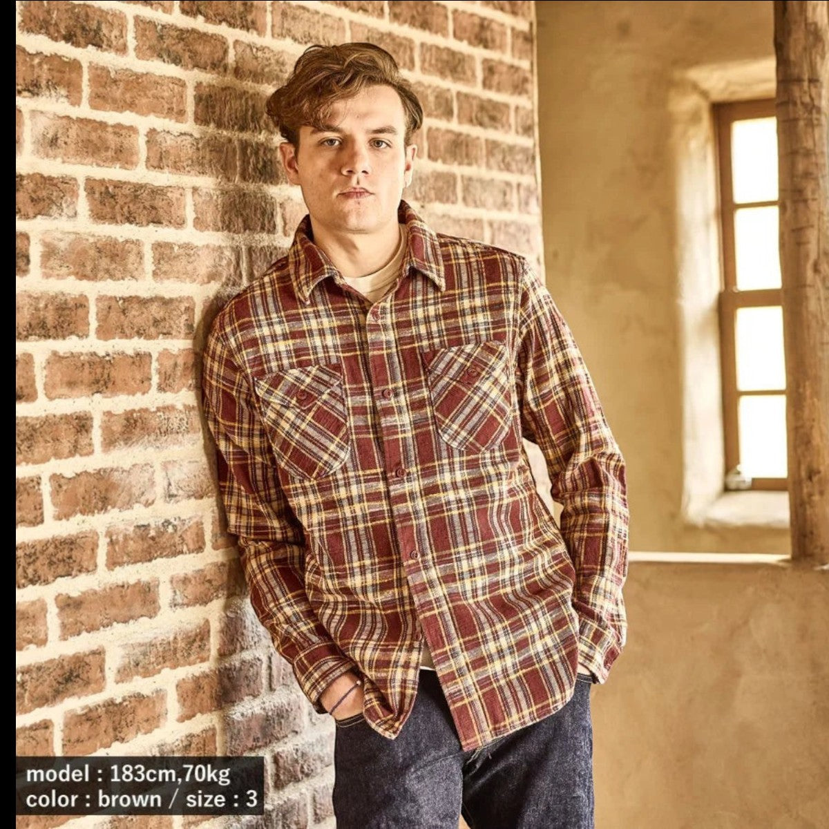 A model wearing a MILWORKS brown slub heavy flannel shirt with a small check pattern, leaning against a brick wall.