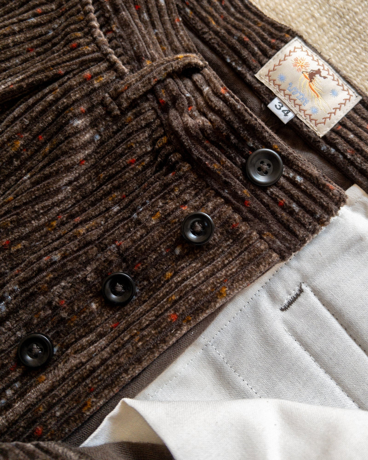 MILWORKS Donegal corduroy pleated chinos in rustic brown with wide wale texture, button fly, and interior label.