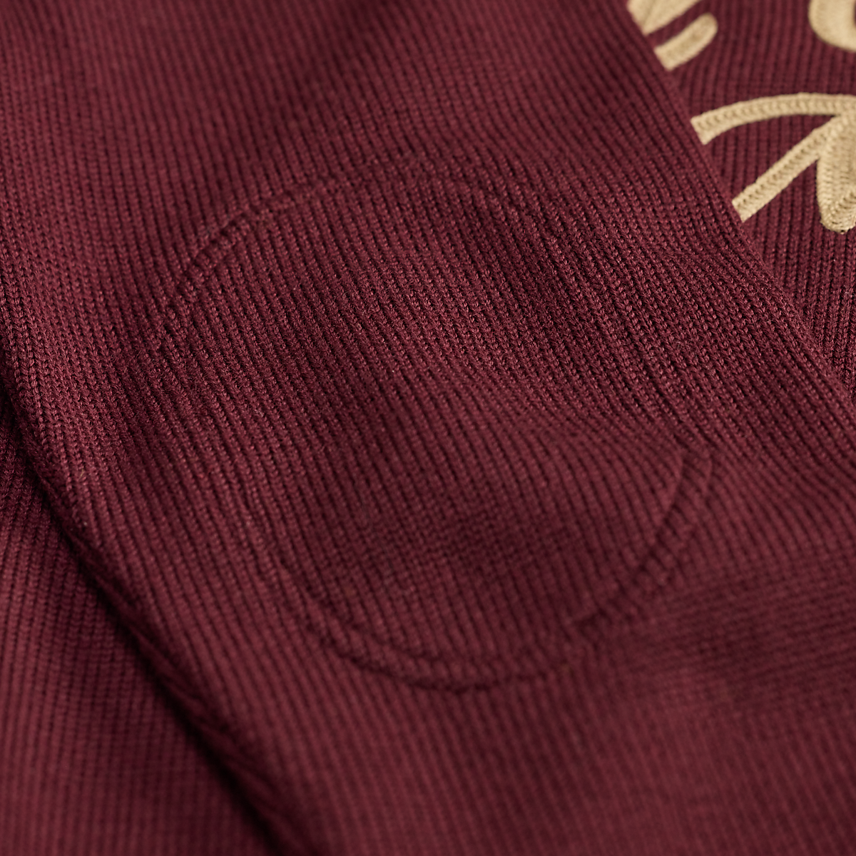 Close-up of MILWORKS maroon wool sweater featuring a textured elbow patch and cream script embroidery on the chest.