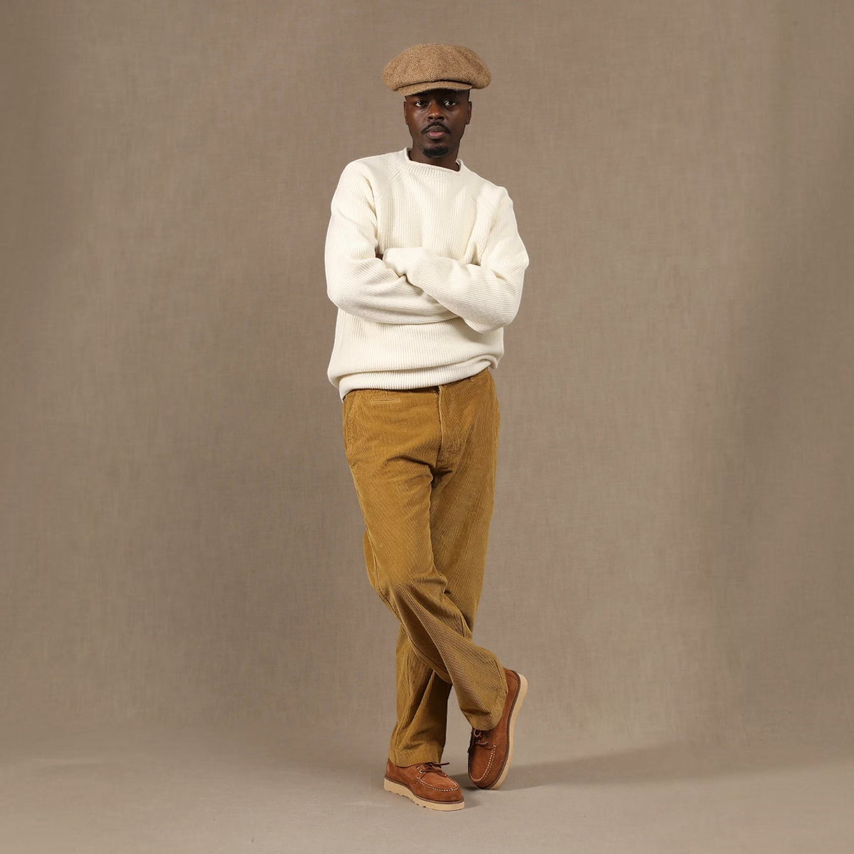 MILWORKS model wearing mustard cotton corduroy Axel chinos, white ribbed sweater, and a brown flat cap.
