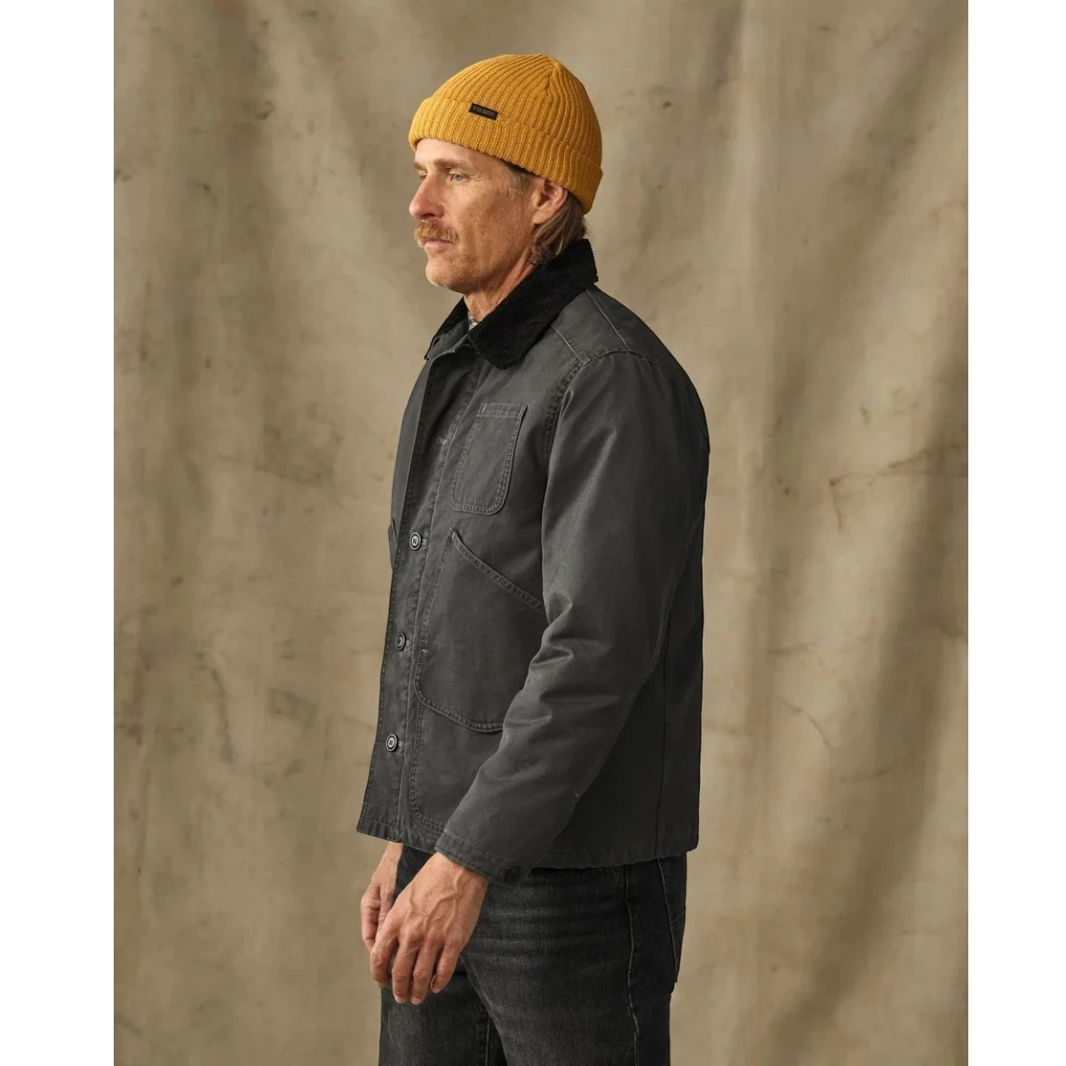MILWORKS model wearing a peat green insulated canvas jacket with a corduroy collar and yellow beanie, side profile