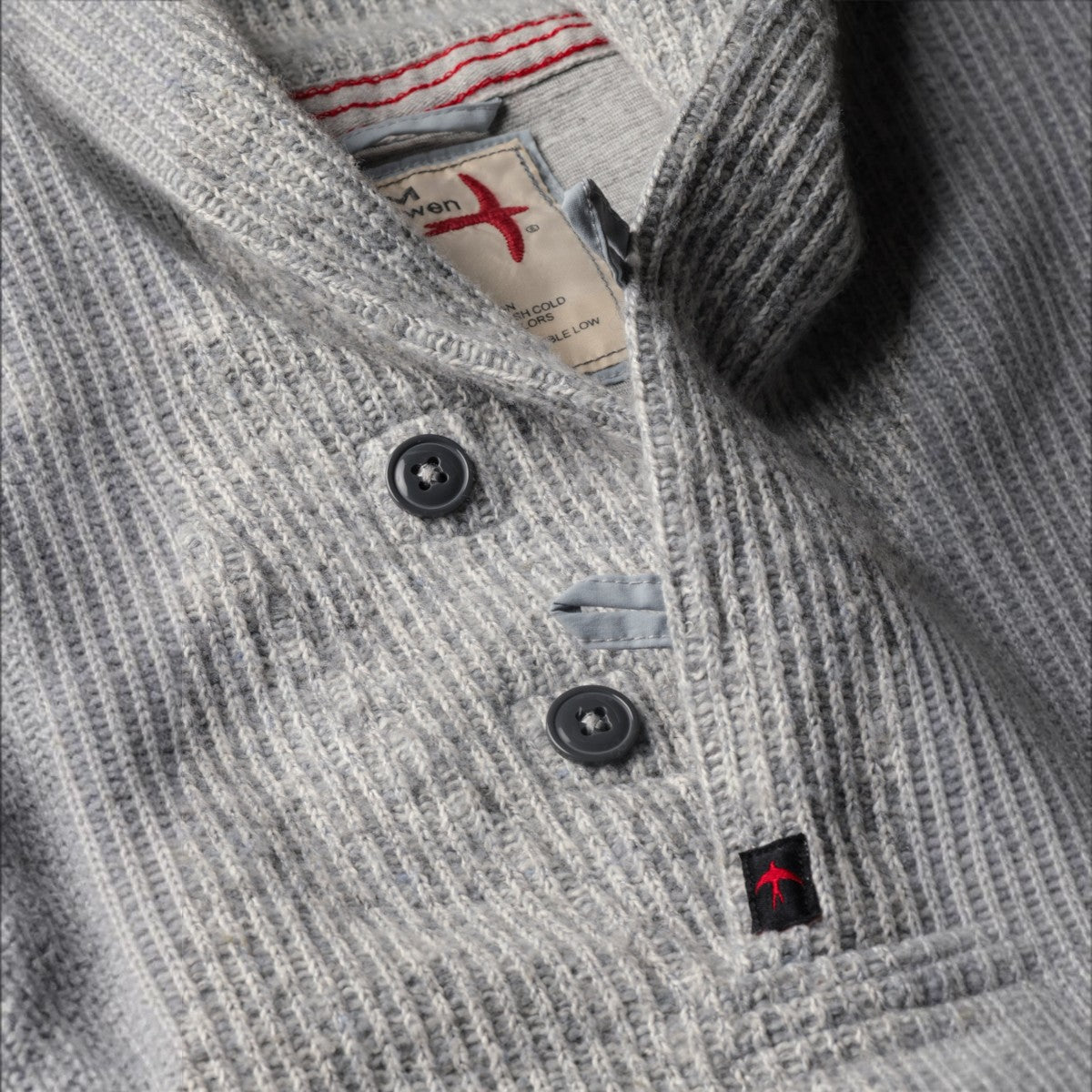 MILWORKS Relwen Ridge Twist Shawl in pale grey featuring a ribbed knit texture, button placket, and red logo detail.