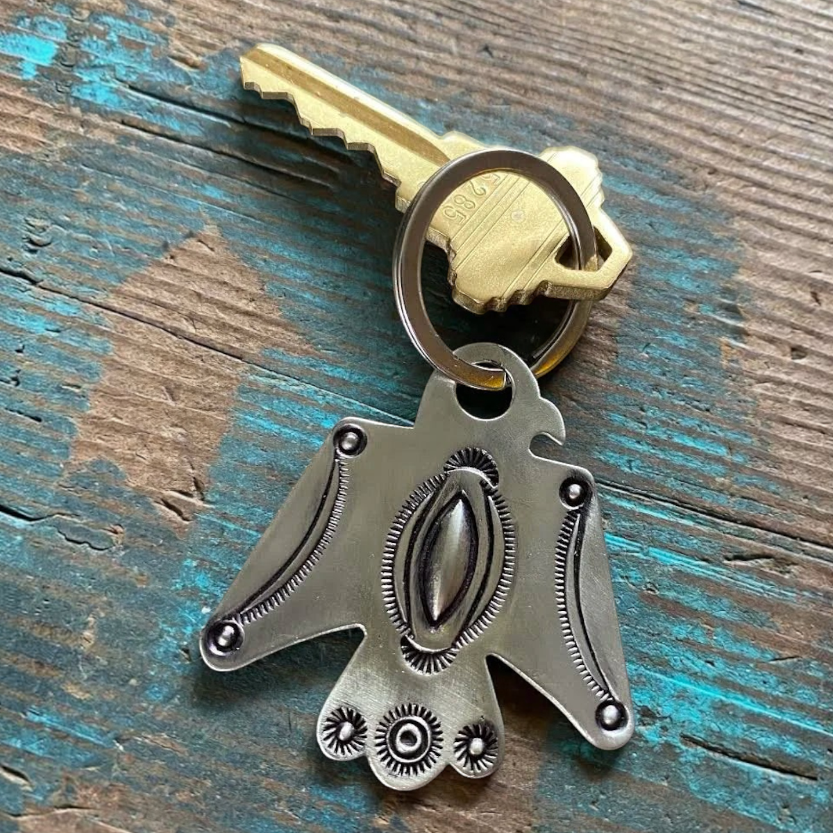 MILWORKS white bronze Thunderbird keychain with repoussé and sunburst stamping details on a rustic wooden surface.
